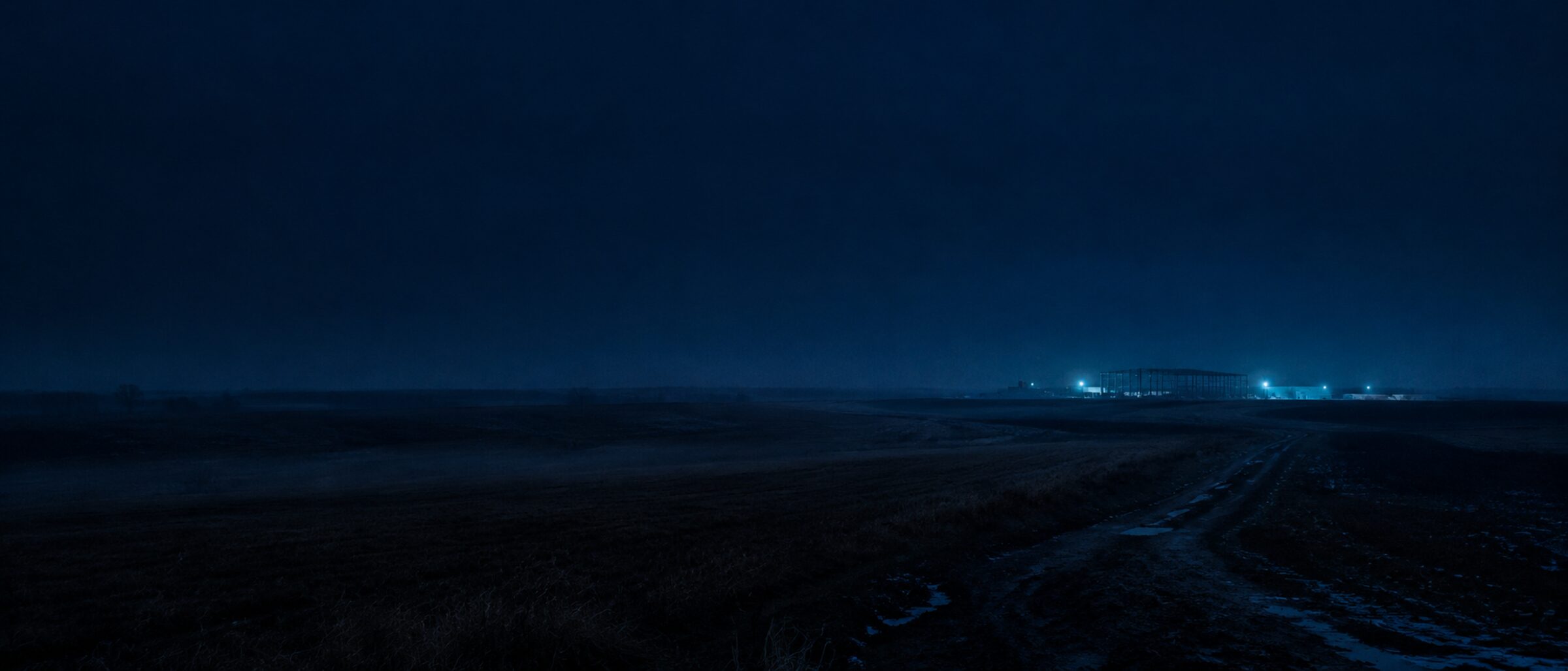 Weite Feldflur, ein Feldweg führt zu einem einzelnen Rohbau am Horizont